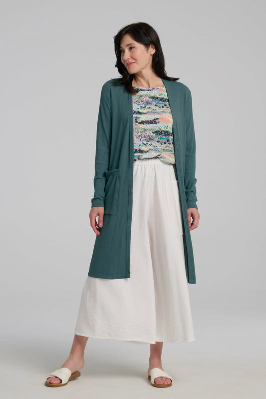 A woman wearing the Streliski Cardigan from Kollontai in Teal, a long open cardigan in bamboo rib with large patch pockets. She is wearing it with a patterned top, and white pants on a plain background