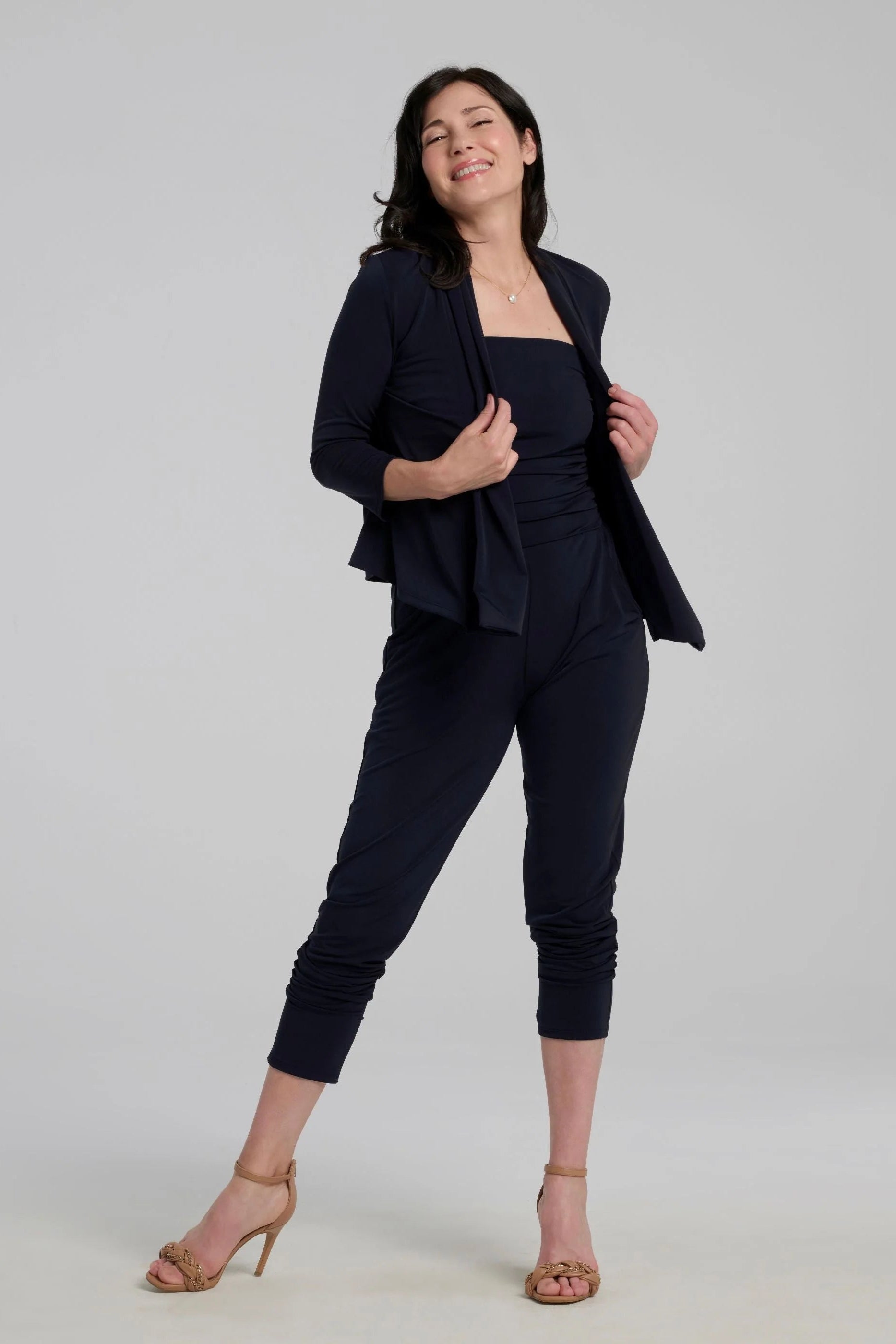 A woman wearing the Lioren Cardigan by Kollontai in Navy, a stretchy cropped jacket with longer pointed panels in the front that can be worn loose or knotted. She is wearing it with  a jumpsuit and standing in front of a grey background 