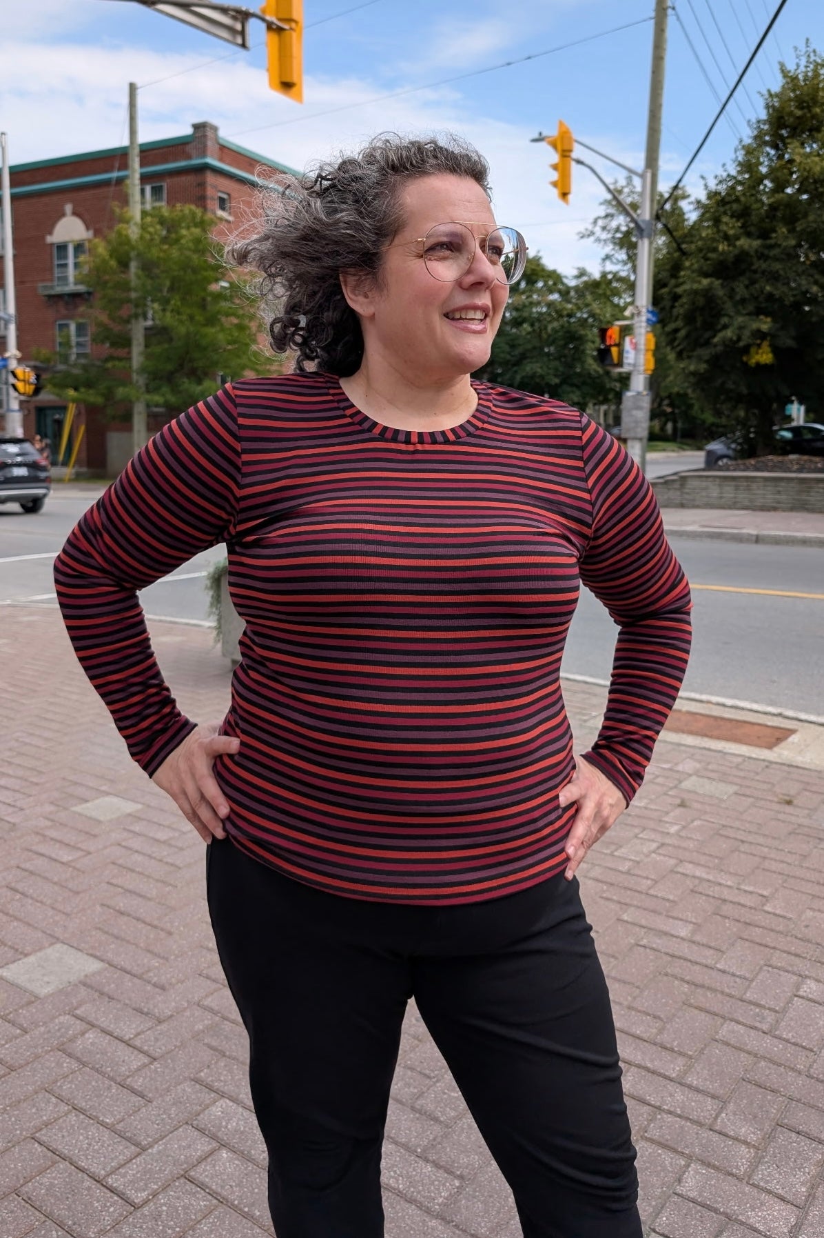 A woman wearing the A25 Sweater by Slak in Colourful Stripe, a long sleeved top with a round neck and a slim fit. She is wearing it with black pants and standing on the street. 