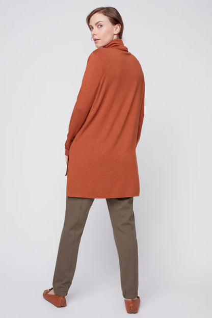 Back view of a woman wearing the Melanie Tunic by Julei in Ochre, a long sleeved turtleneck tunic with a hi-low hemline and side slits. She is wearing it with green pants and standing in front of a white background. 