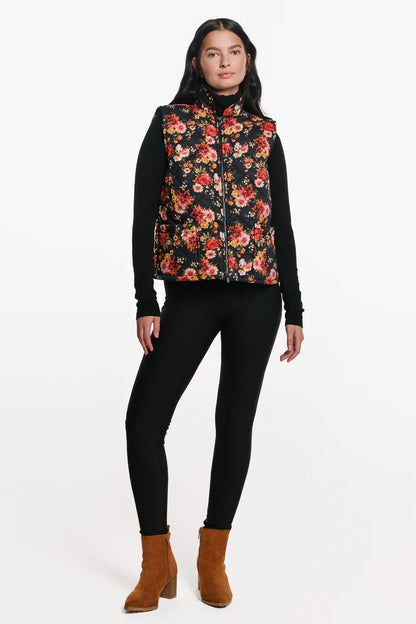 A woman wearing the Harper Vest by EMK, a quilted floral vest with a zip front two pockets, and black waffle knit fabric at the collar. She is wearing it with black pants and standing in front of a white background. 
