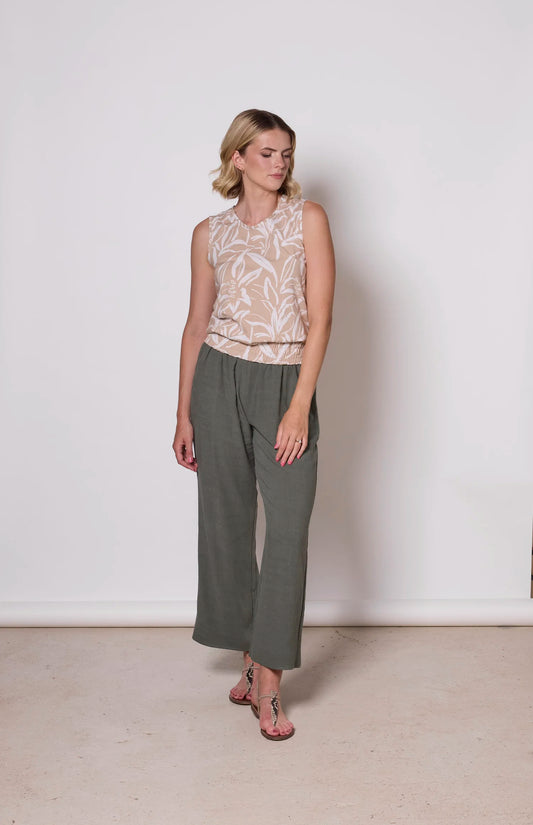 A woman wearing the Heidi Pants by Tangente in Thyme, wide legged slightly cropped pants with an elastic waist. She is wearing them with a printed beige tank top and standing in front of a white background. 