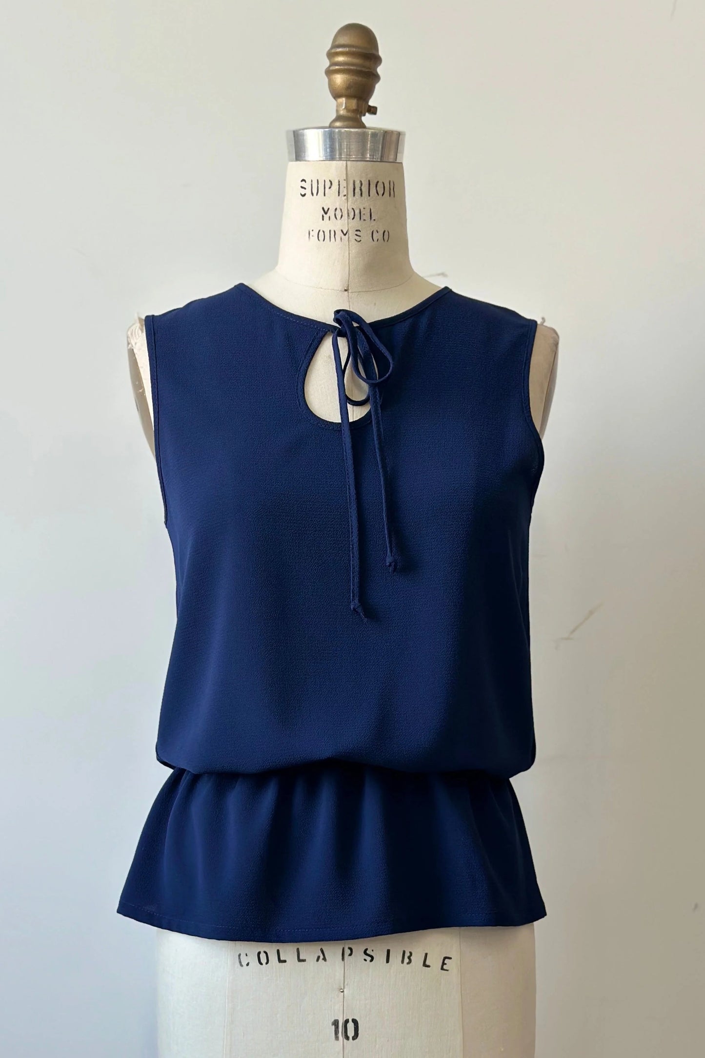 The Serena Top from Kollontai in Navy, a sleeveless top with a keyhole and tie neckline, elastic waist and flared peplum hem, is shown on a mannequin against a white background. 