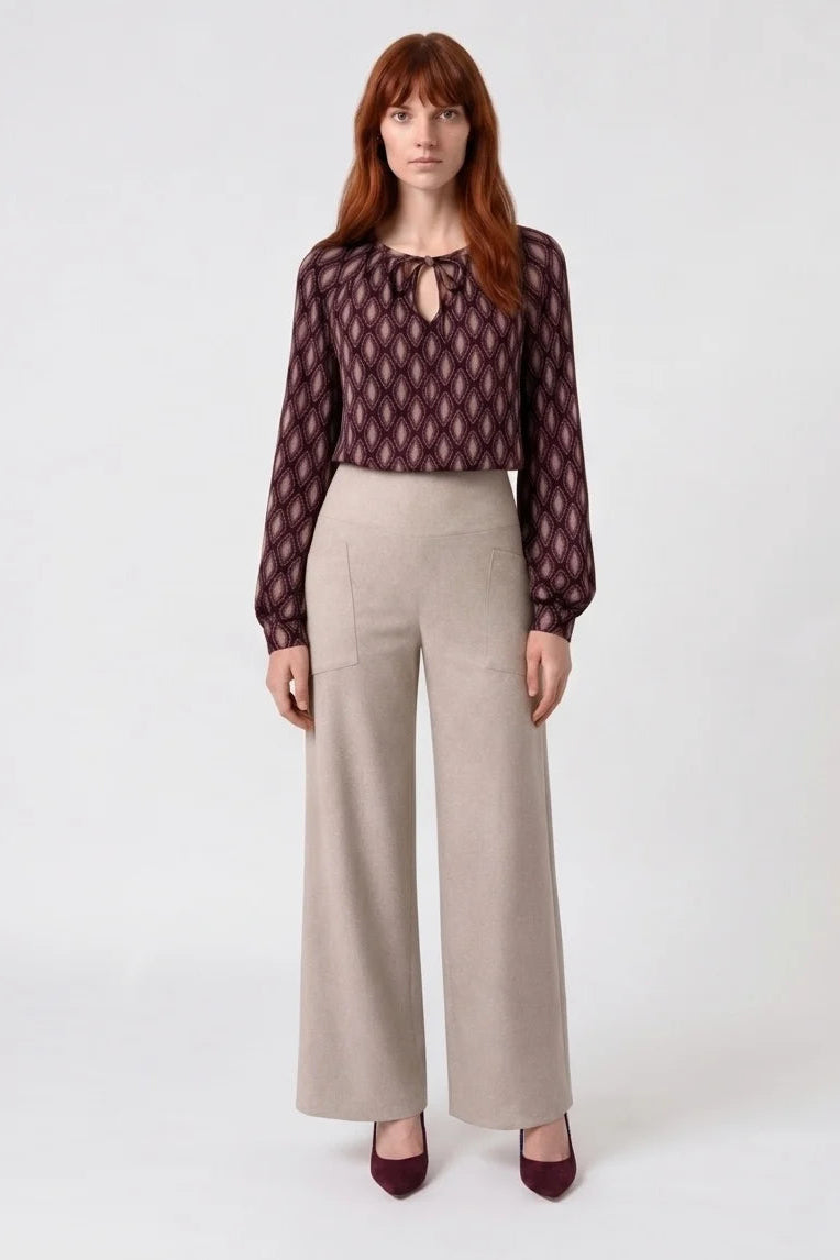A woman wearing the Isabella Blouse by Kollontai, a burgundy and pink patterned blouse with a tie detail at the neck, balloon sleeves with square jewel buttons, and an elastic waist. She is wearing it with beige pants and standing in front of a white background. 