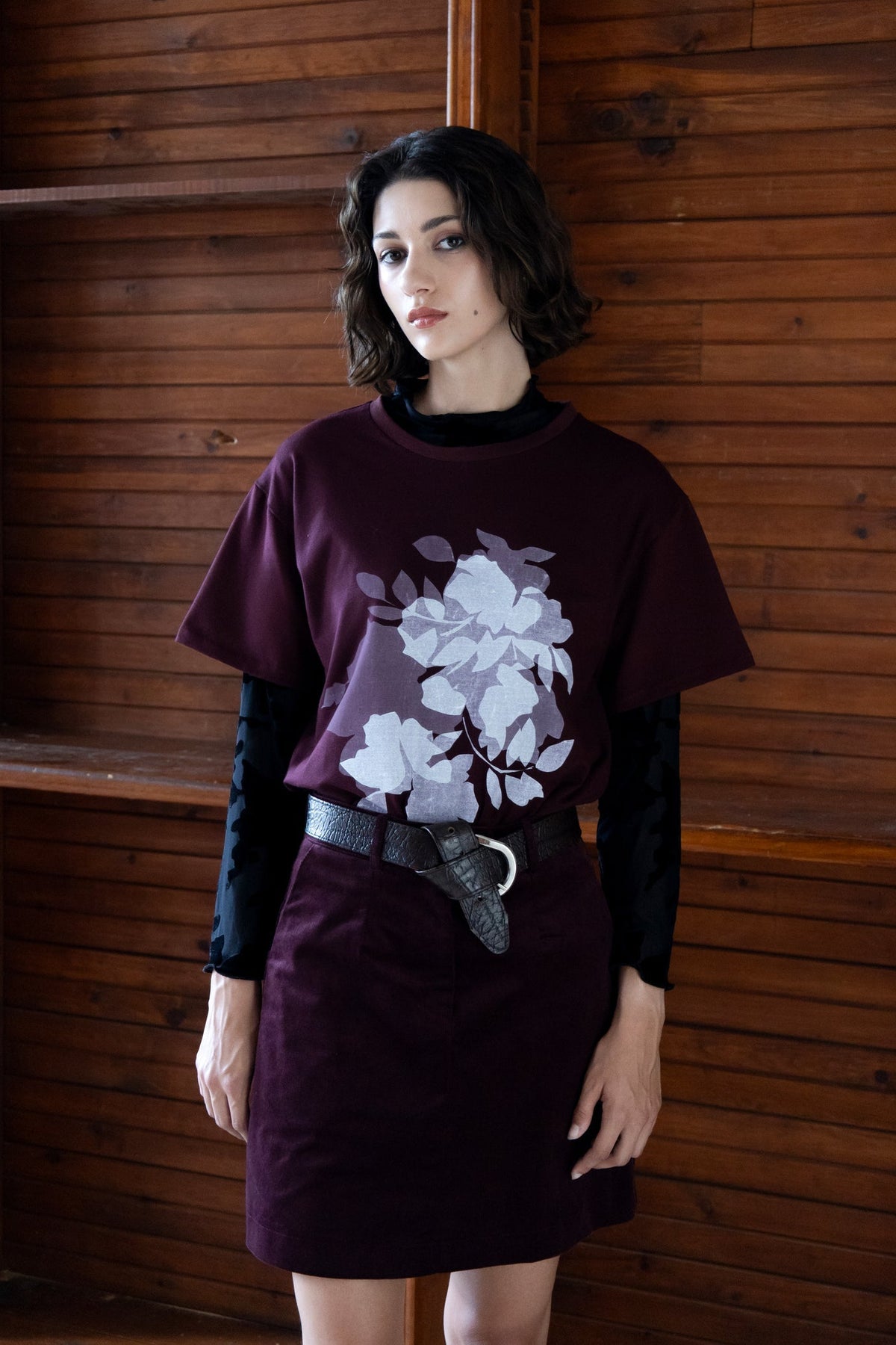 A woman wearing the Tiger's Eye Screen-printed T-shirt by Eve Lavoie, an oversized Burgundy t-shirt with a white graphic print on the front. She is wearing it with a long sleeved black shirt underneath and a burgundy miniskirt and standing indoors by a wooden wall. 