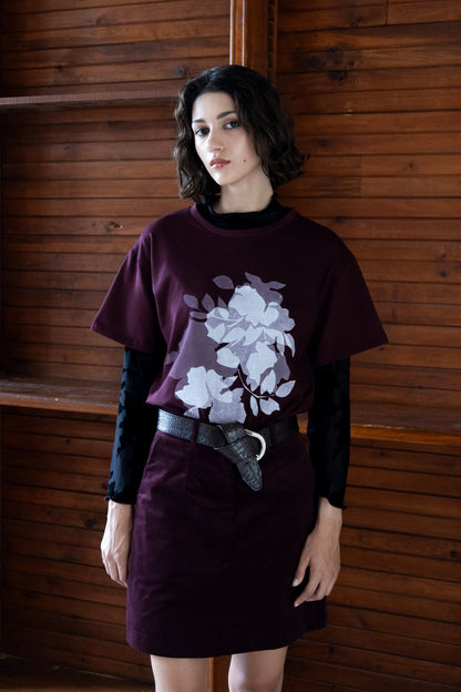 A woman wearing the Tiger's Eye Screen-printed T-shirt by Eve Lavoie, an oversized Burgundy t-shirt with a white graphic print on the front. She is wearing it with a long sleeved black shirt underneath and a burgundy miniskirt and standing indoors by a wooden wall. 