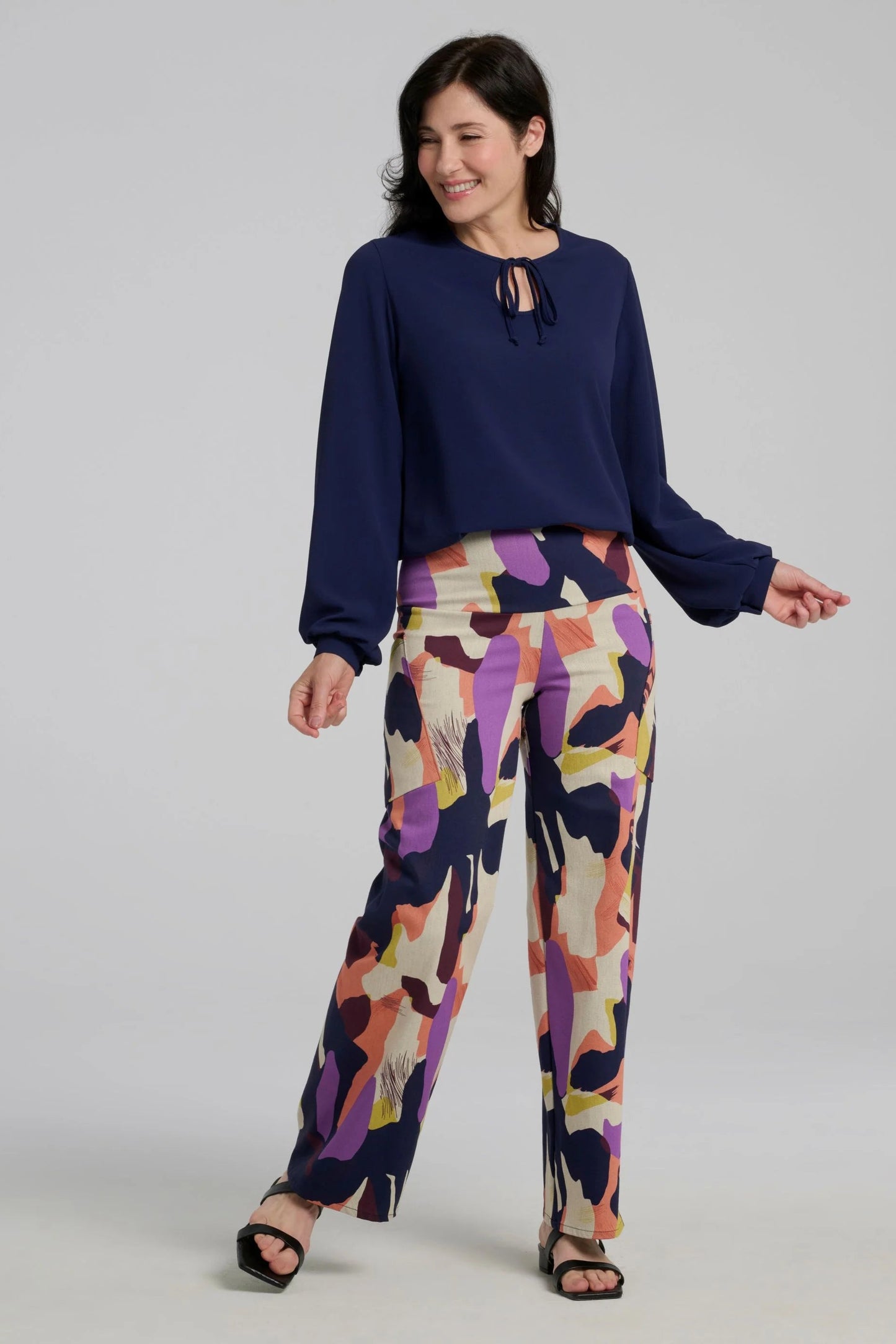 A woman wearing the Elma Pants by Kollontai in a Multicolour abstract print, with a wide pull-one waistband, straight legs, and slanted pockets. She is wearing them with a navy top and 
and standing in front of a plain background