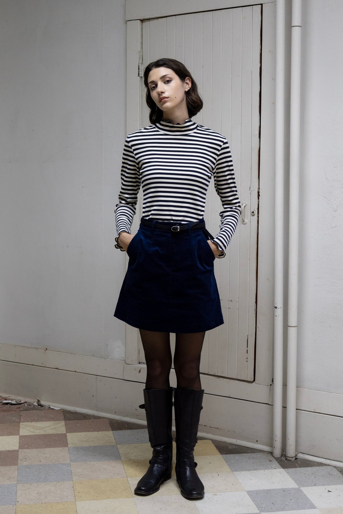 A woman wearing the Onyx Mini Skirt by Eve Lavoie in Navy, a corduroy mini with a zipper, oversized button and back slit. She is standing in a room with a checkered floor and white walls, and wearing it with a striped turtleneck. 