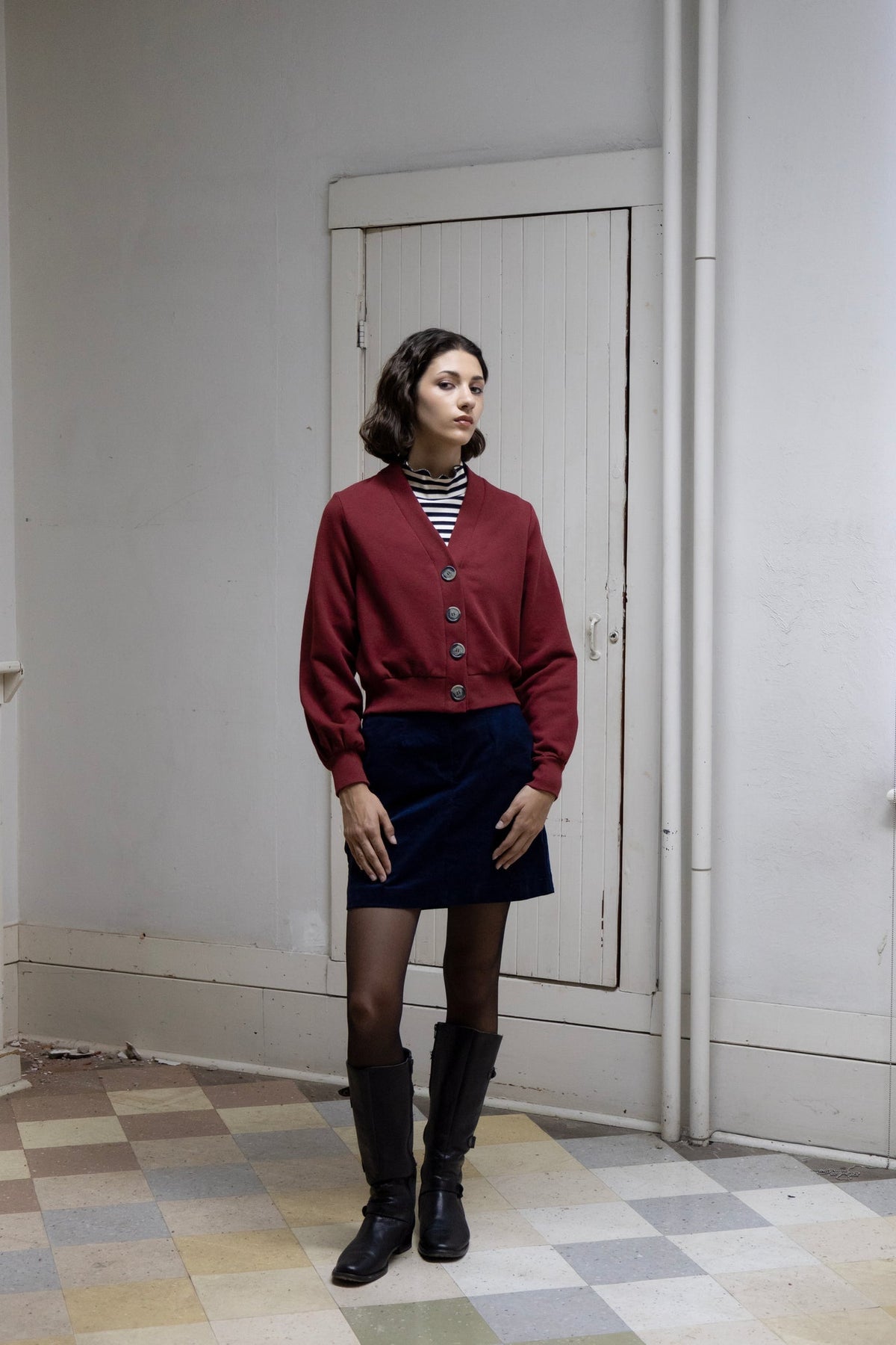 A woman wearing the Onyx Mini Skirt by Eve Lavoie in Navy, a corduroy mini with a zipper, oversized button and back slit. She is standing in a room with a checkered floor and white walls, and wearing it with a burgundy cardigan over a striped turtleneck. 