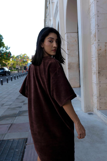 A woman wearing the Mykonos Dress by MAS in Cacao, a short sleeved boxy, knee-length dress in ribbed velvet fabric. She is walking down a city street. 