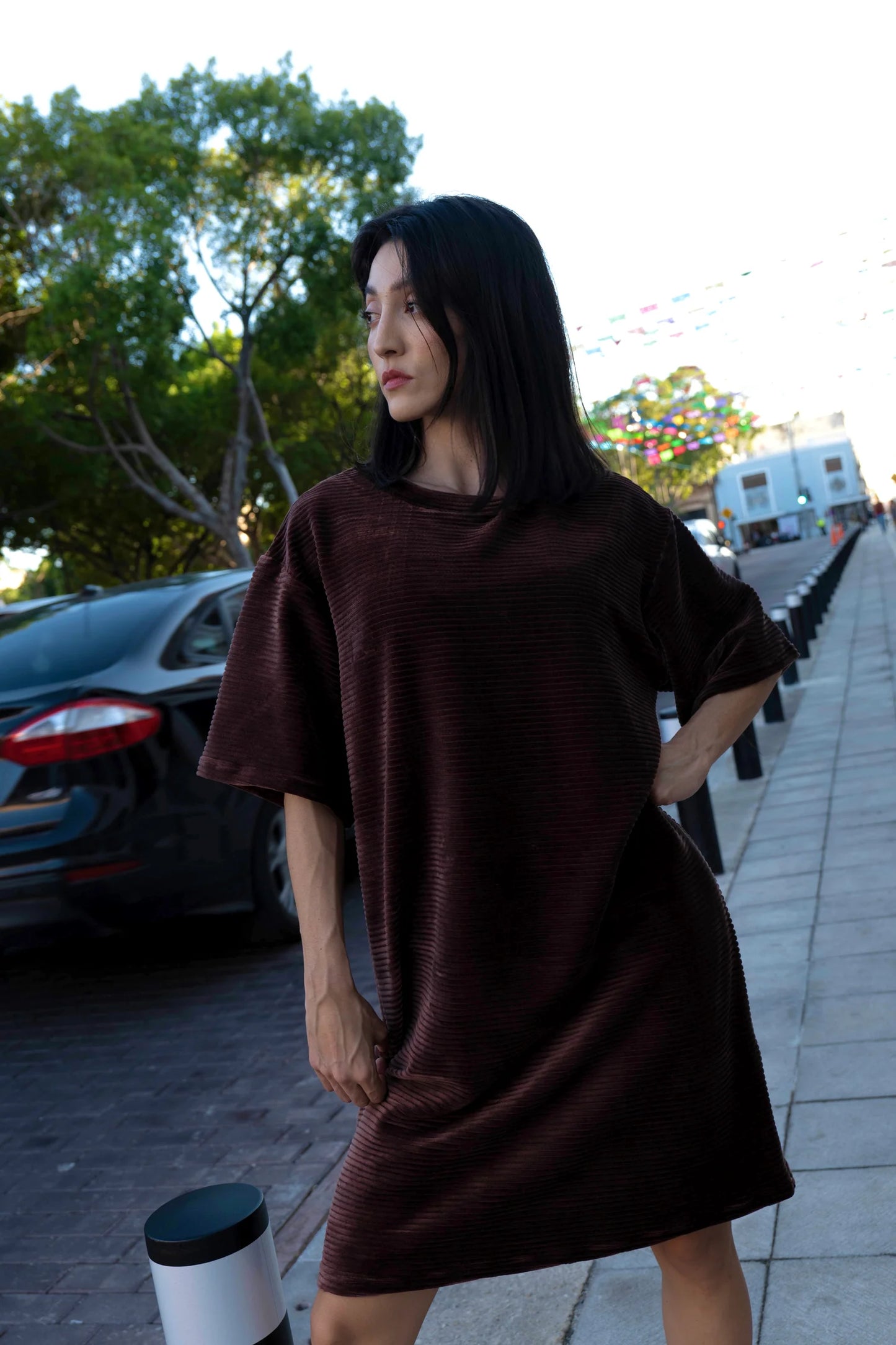 A woman wearing the Mykonos Dress by MAS in Cacao a short sleeved boxy, knee-length dress in ribbed velvet fabric. She is standing on a city street.