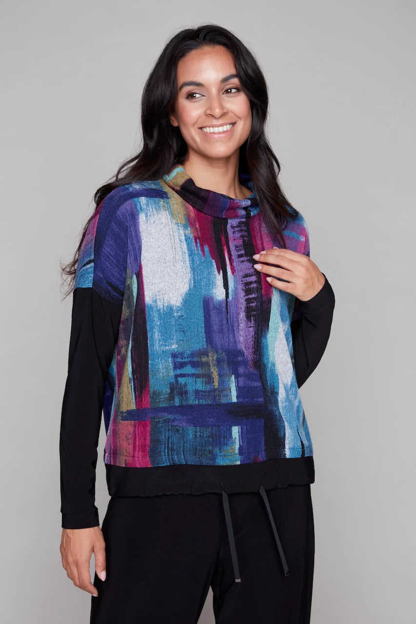 A woman wearing the Payton Top by Compli K in Blue and Berry Abstract print, with a turtleneck, drop sleeves, and drawstring waist. She is wearing it with black pants and standing in front of a grey background. 