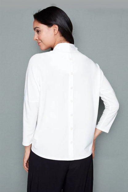 Back view of a woman wearing the Perry Top by Compli K in Ivory, featuring a cowl neck, 3/4 drop sleeves, and decorative buttons up the back. She is wearing it with black pants and standing in front of a white background. 