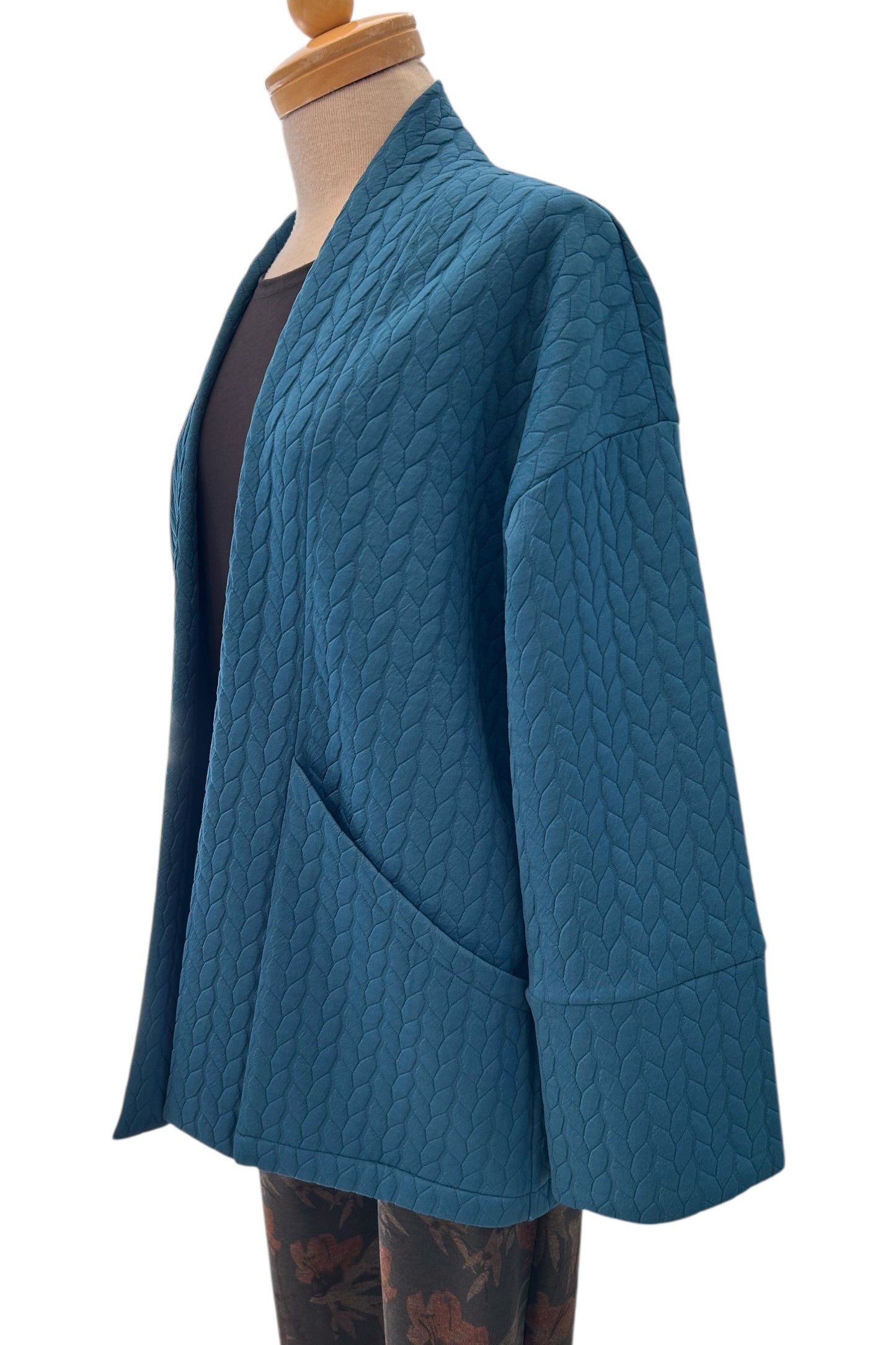 The Ina Coatigan by Tangente in Peacock, an open front cardigan in a cable-knit textured fabric, with dropped sleeves and patch pockets, is shown on a mannequin against a white background. 