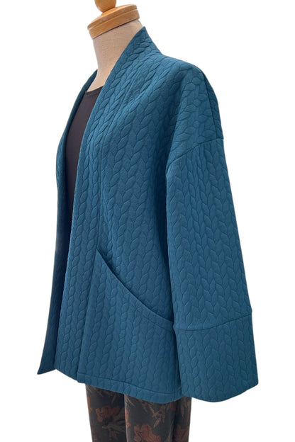 The Ina Coatigan by Tangente in Peacock, an open front cardigan in a cable-knit textured fabric, with dropped sleeves and patch pockets, is shown on a mannequin against a white background. 