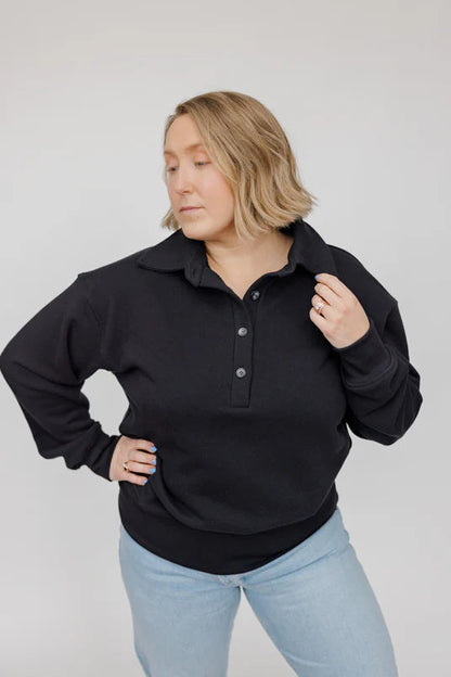 A woman wearing the Classic Coastal Sweater by Blondie in Black, a relaxed fit sweatshirt with a collar and four button placket. She is wearing it with jeans and standing in front of a white background. 