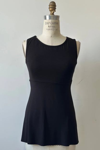 The Nira Top by Kollontai in Black, a sleeveless top with a wide neck, an empire waist, and a cut that's fitted through the bodice and flares away from the torso, is shown on a mannequin against a white background. 