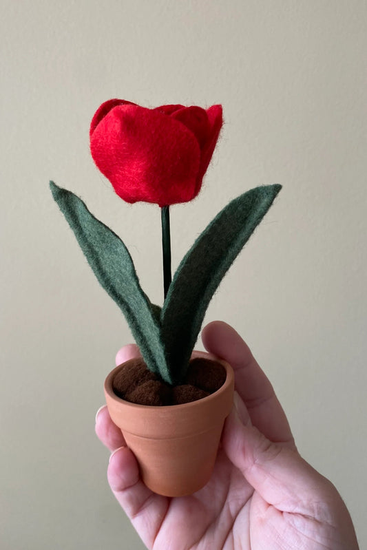A hand holding up the Handmade Felt Tulip by ForgetBKnot is shown against a beige background. 