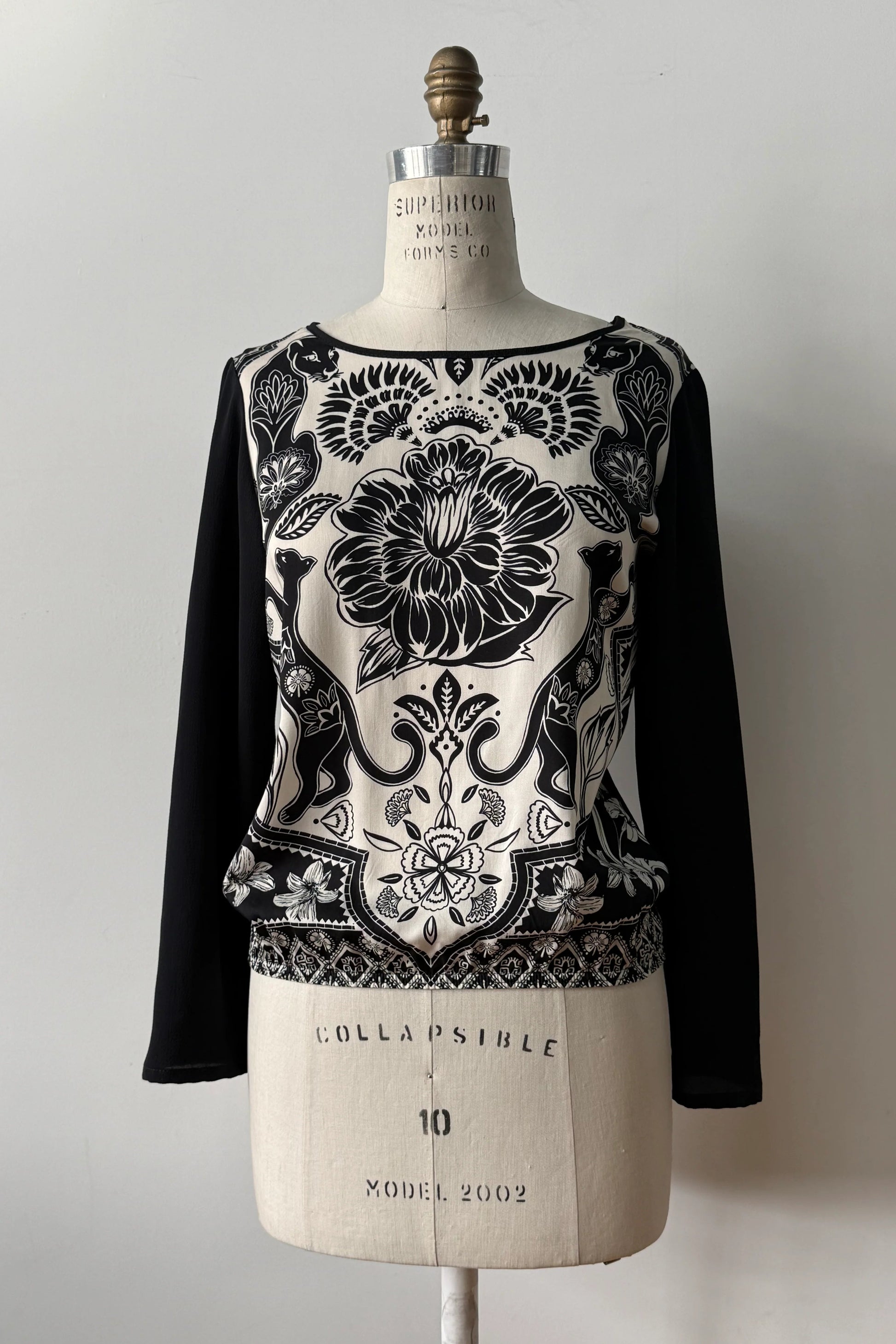 The Oriane Blouse by Kollontai, featuring a black-on-ivory floral and panther print, solid black long sleeves and back, and an elasticized hem, is shown on a mannequin against a grey background. 