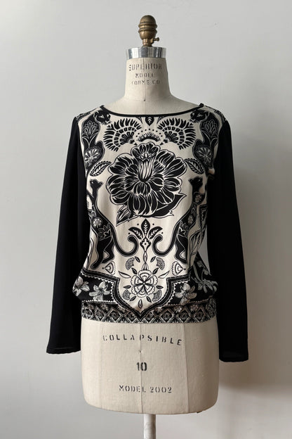 The Oriane Blouse by Kollontai, featuring a black-on-ivory floral and panther print, solid black long sleeves and back, and an elasticized hem, is shown on a mannequin against a grey background. 