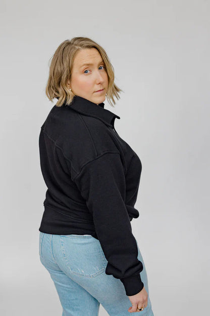 Back view of a woman wearing the Classic Coastal Sweater by Blondie in Black, a relaxed fit sweatshirt with a collar and four button placket. She is wearing it with jeans and standing in front of a white background. 