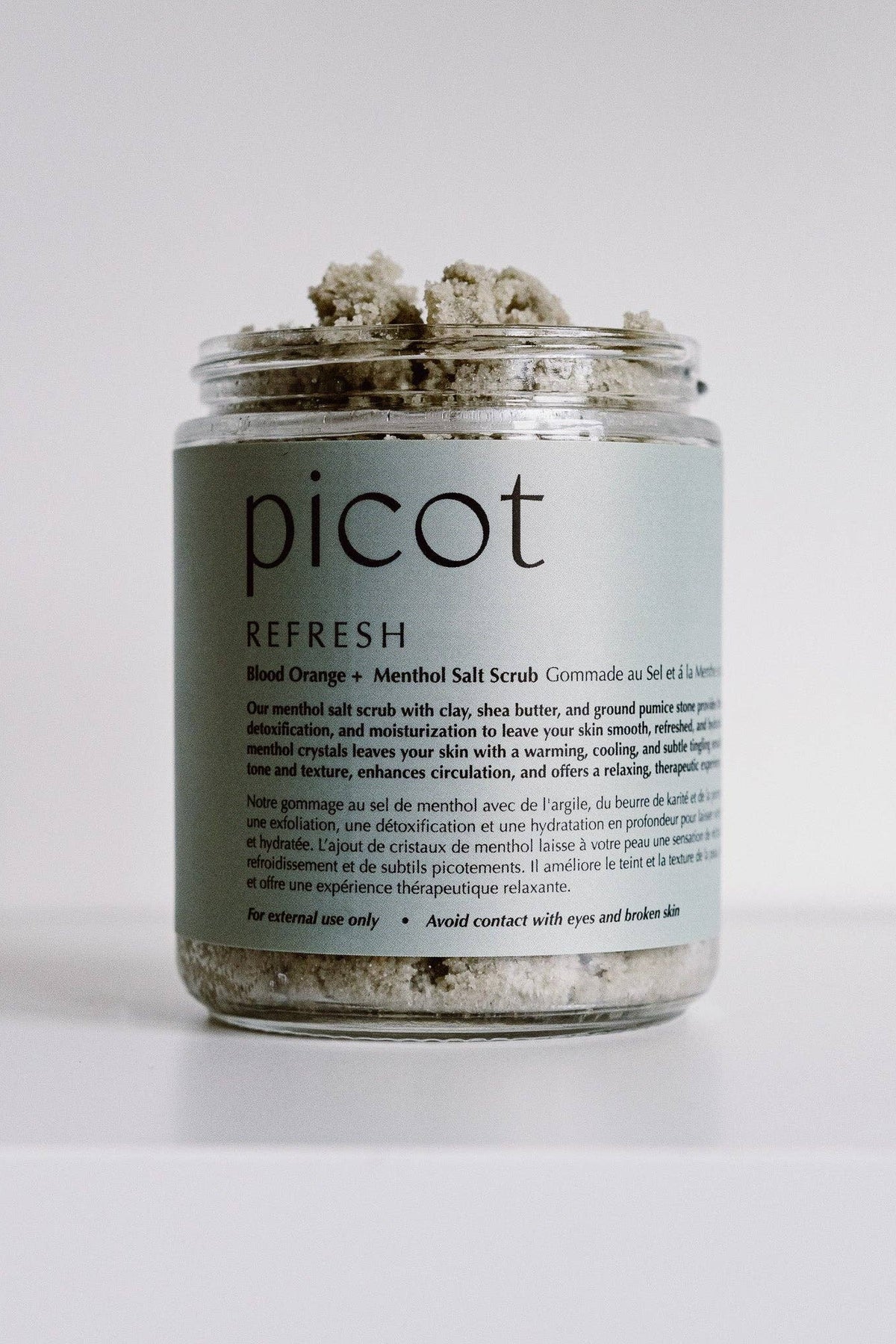 A clear glass jar of Picot Refresh Blend Orange + Menthol Salt Scrub containing textured herbal scrub, set against a plain white background.