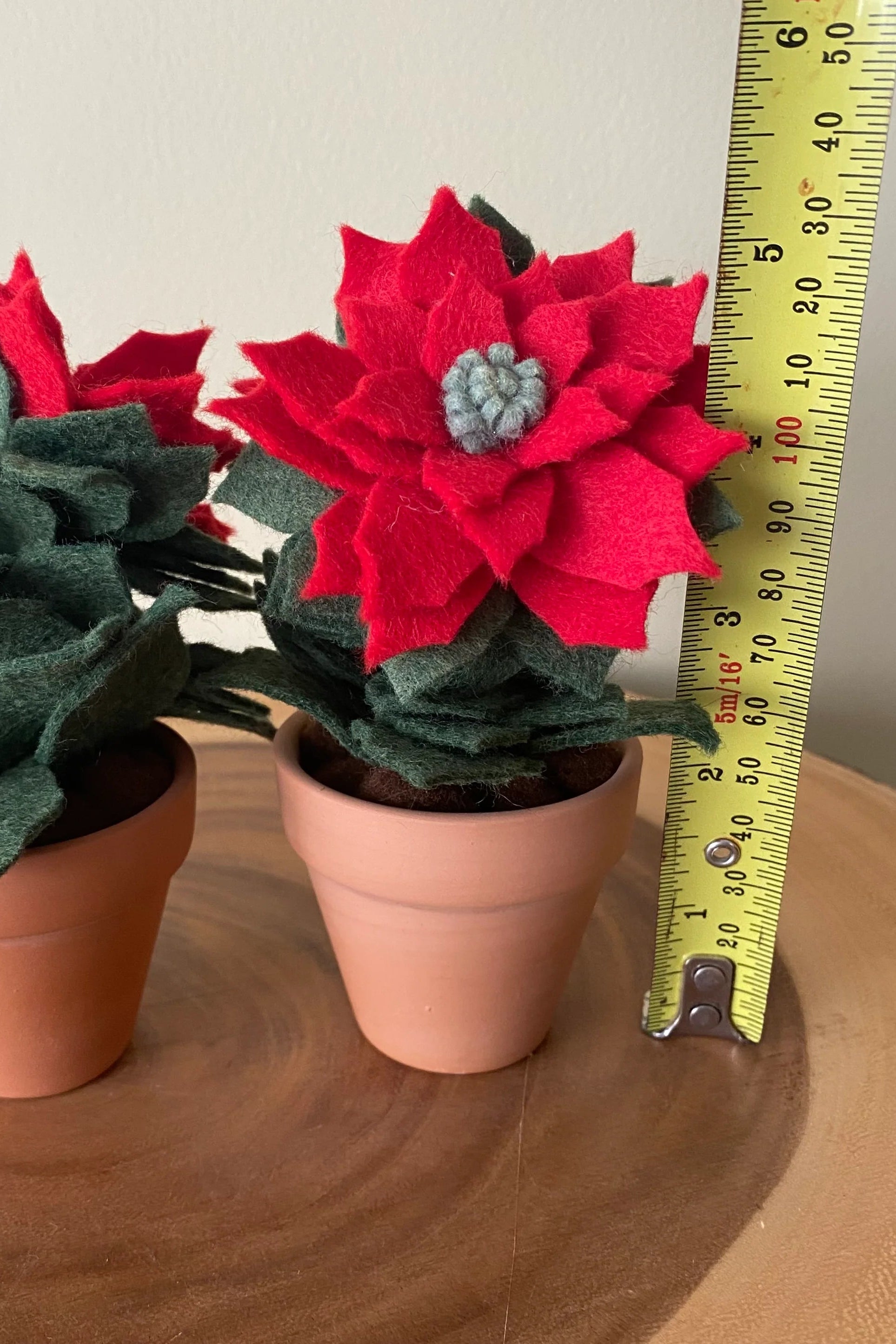 The Holiday Felt Poinsettia by ForgetBKnot is shown with a ruler for scale on a wooden surface.