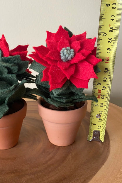 The Holiday Felt Poinsettia by ForgetBKnot is shown with a ruler for scale on a wooden surface.
