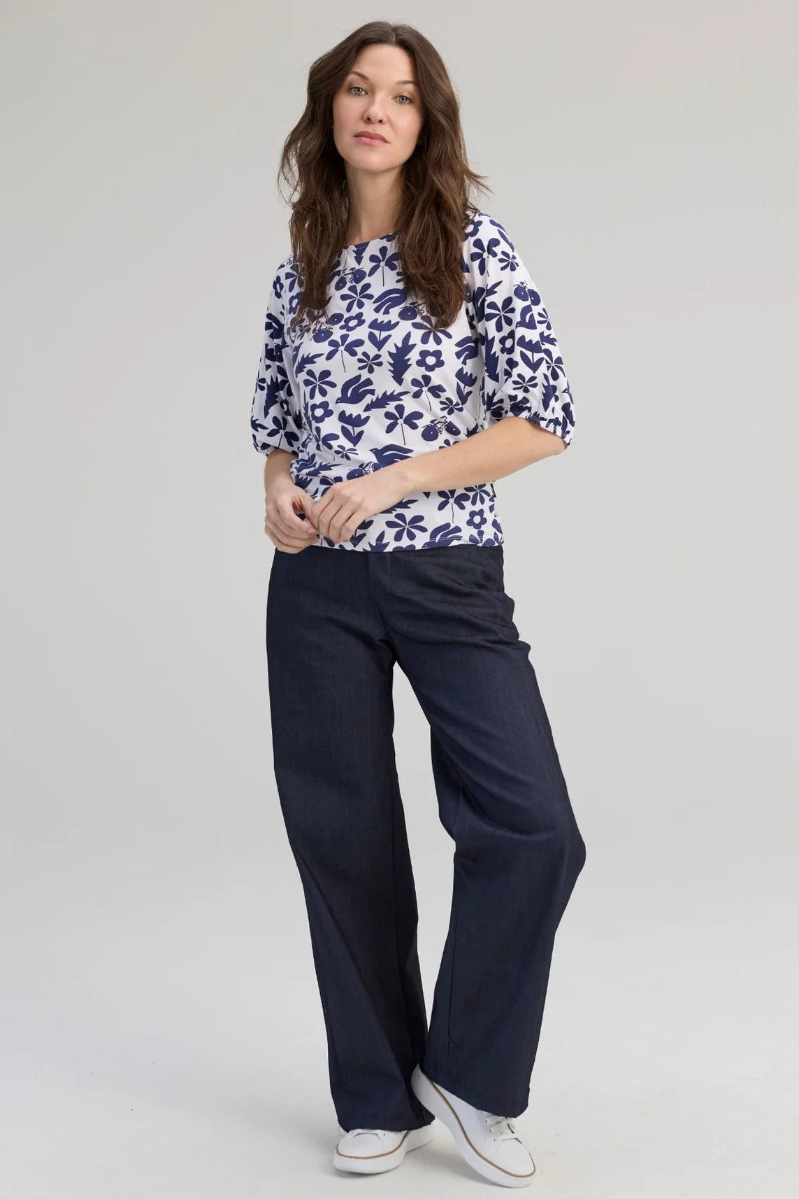A woman wearing the Cadieux Top from Message Factory in a floral Navy Print, with 3/4 sleeves with gathered cuffs, a wide neck and slim fit.  She is wearing it with dark pants and standing against a plain background. 