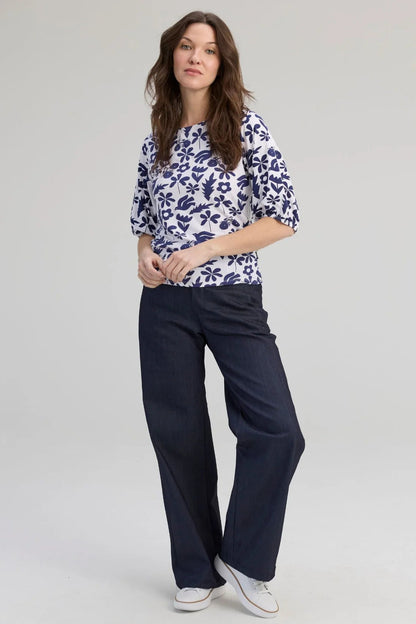 A woman wearing the Cadieux Top from Message Factory in a floral Navy Print, with 3/4 sleeves with gathered cuffs, a wide neck and slim fit.  She is wearing it with dark pants and standing against a plain background. 