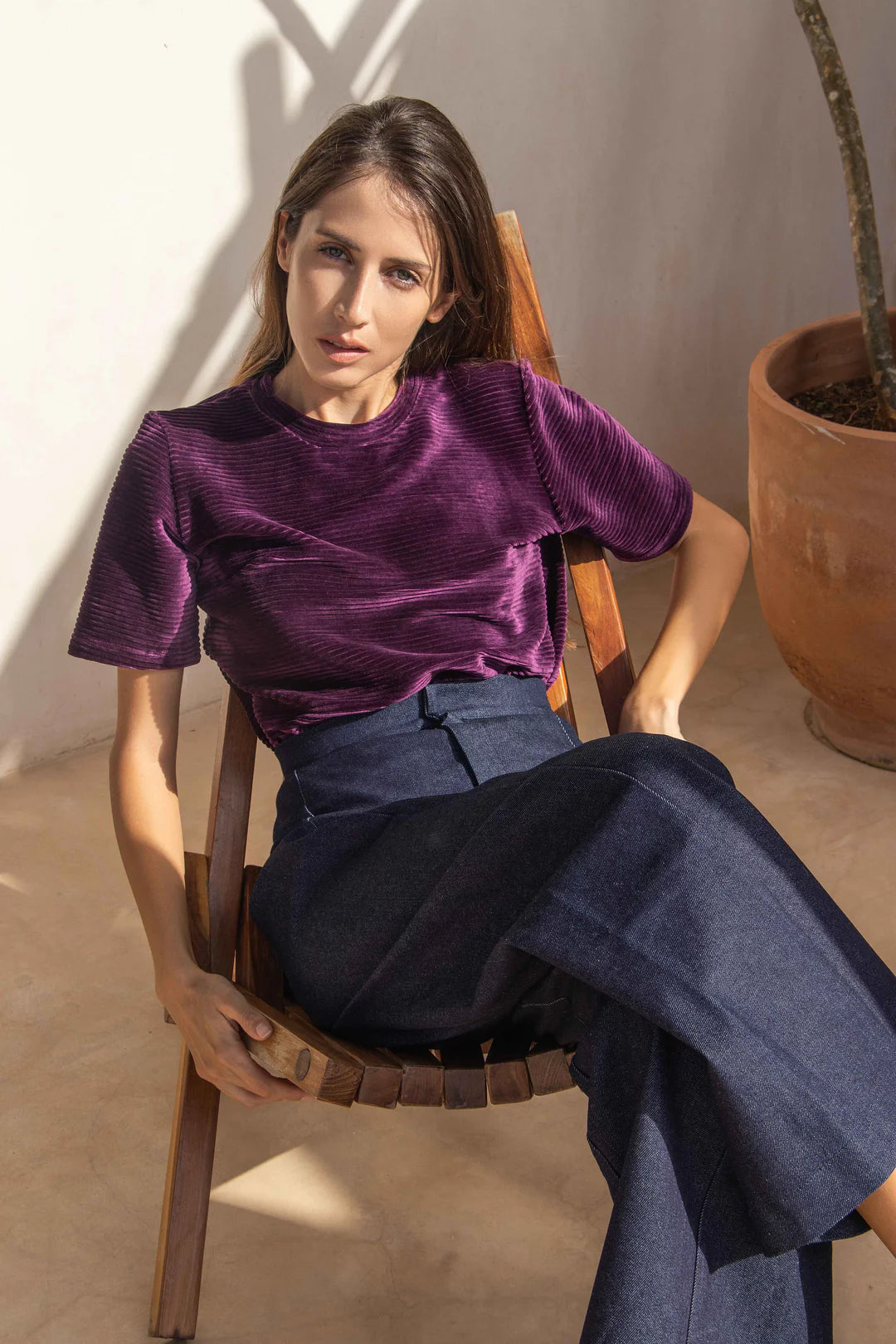 A woman wearing the Vienna Pants by MAS in Jeans, with a wide leg, side seams that sit slightly to the front, and two back pockets, with the Tofino Top in Amethyst. She is sitting in a wooden chair. 