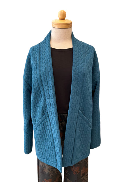 The Ina Coatigan by Tangente in Peacock, an open front cardigan in a cable-knit textured fabric, with dropped sleeves and patch pockets, is shown on a mannequin against a white background. 