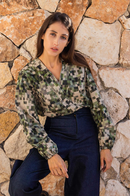 A woman wearing the Manama Blouse by MAS in a pixellated Camo Print, featuring a modest cross-over neckline, darts, and long sleeves with elastic cuffs, with the Vienna Pants in Jeans. She is standing in front of a stone wall. 