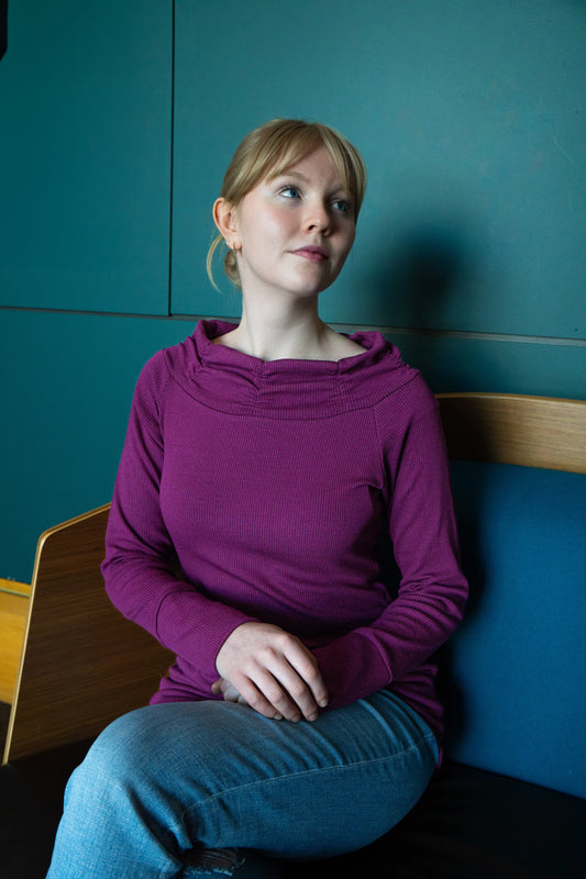 A woman wearing the Myrtille Sweater by Karkass in Purple, with a large pleated cowl neck, long sleeves, and wide band at the bottom. She is wearing it with jeans and sitting against a teal wall.