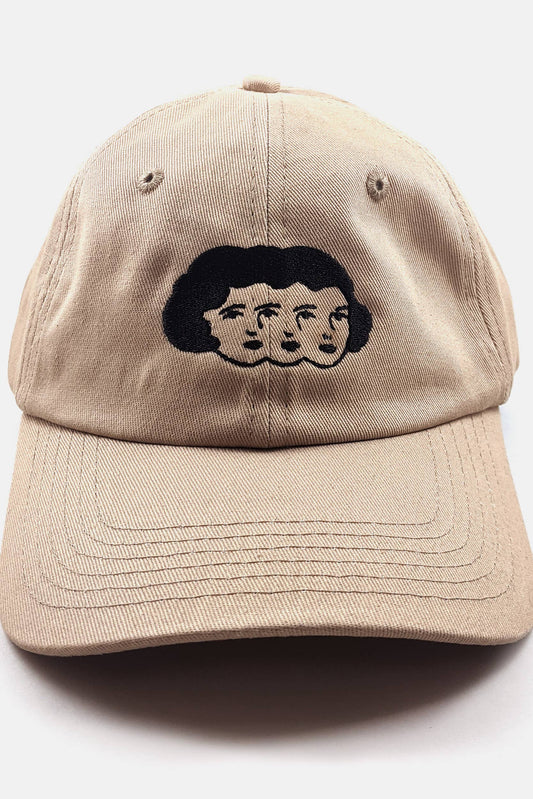 The Triplets Hat from Badaboöm Studio, featuring embroidery of a woman's face in triplicate in black on a tan background, is shown against a light grey background. 