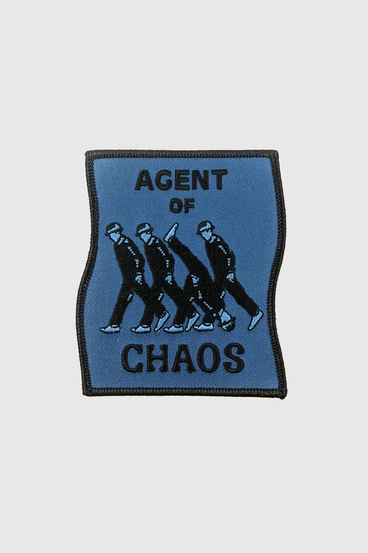 The Agent of Chaos patch from Badaboöm Studio, a rectangular blue patch depicting three men walking upright and one walking upside down, with the words of Agent of Chaos in black text, is shown against a white background. 