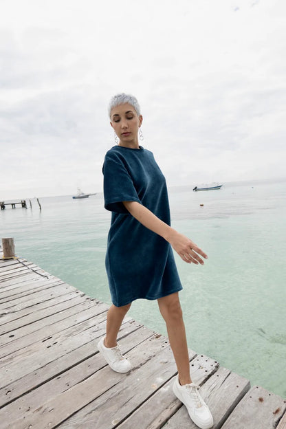 A woman wearing the Mykonos Dress by MAS in Juniper, a short sleeved boxy, knee-length dress in ribbed velvet fabric. She is walking on a wooden dock. 