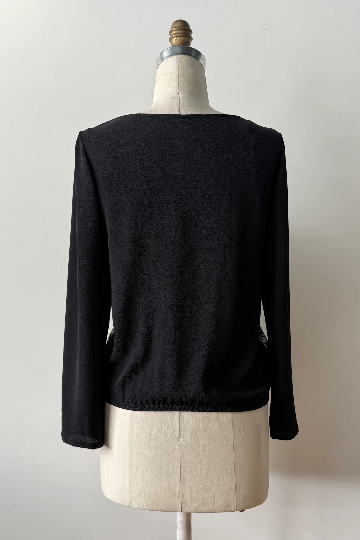 A back view of the Oriane Blouse by Kollontai, featuring a black-on-ivory floral and panther print, solid black long sleeves and back, and an elasticized hem, is shown on a mannequin against a grey background. 