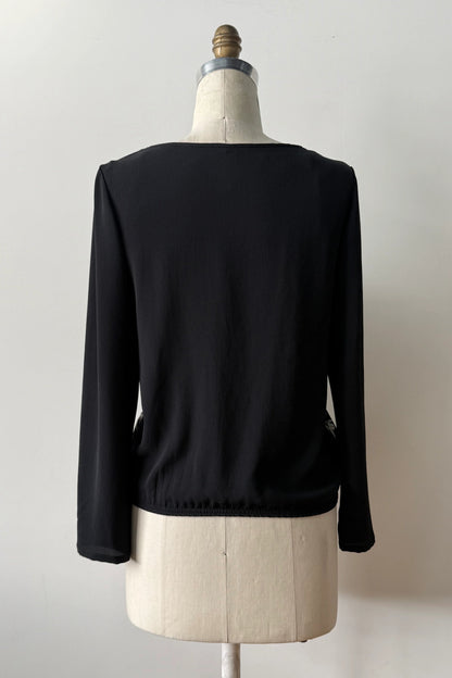 A back view of the Oriane Blouse by Kollontai, featuring a black-on-ivory floral and panther print, solid black long sleeves and back, and an elasticized hem, is shown on a mannequin against a grey background. 