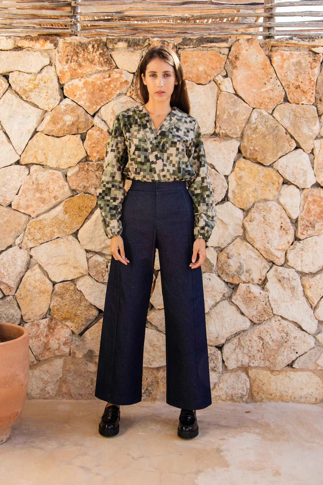 A woman wearing the Manama Blouse by MAS in a pixellated Camo Print, featuring a modest cross-over neckline, darts, and long sleeves with elastic cuffs, with the Vienna Pants in Jeans. She is standing in front of a stone wall. 