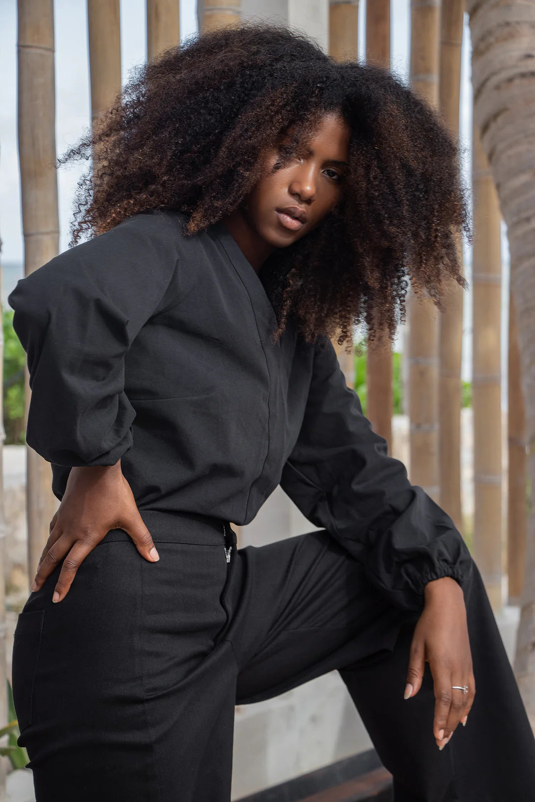 A woman wearing the Vienna Pants by MAS in Pepper, with a wide leg, side seams that sit slightly to the front, and two back pockets, with a black blouse, standing in front of a wooden wall. 