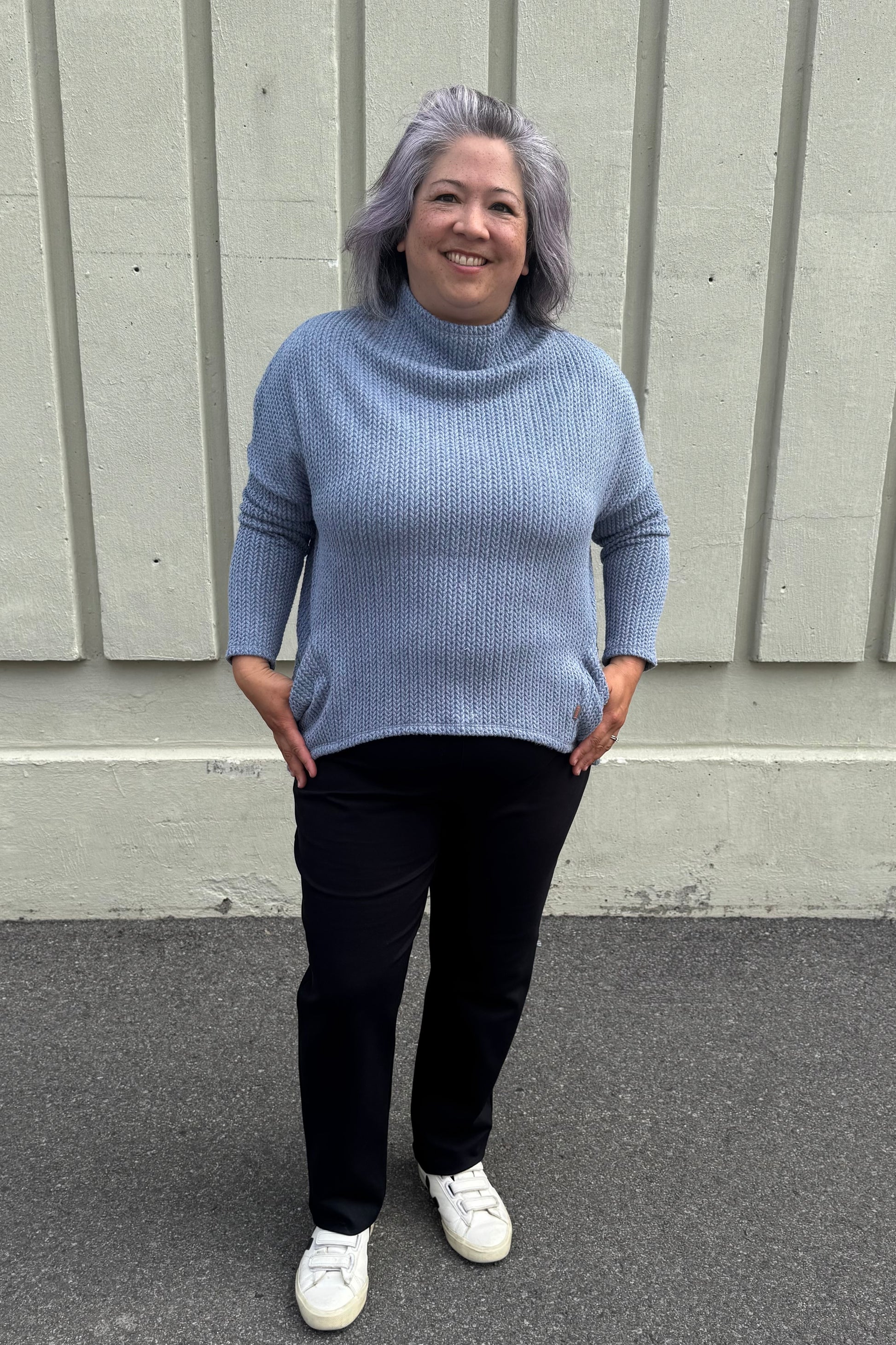 A woman wearing the Meeso Sweater by MOOVment in Denim, a One Size oversized knit sweater with a wide mock turtleneck, long dropped sleeves and large side pockets. She is wearing it with black pants and standing outside in front of a grey wall. 