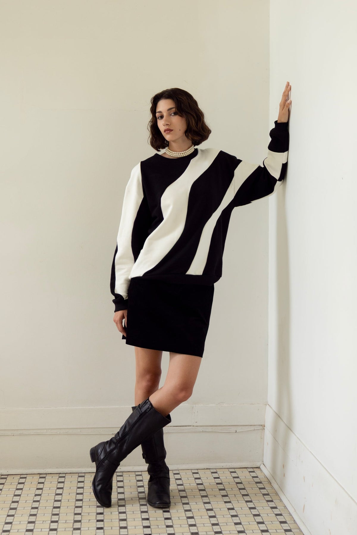 A woman wearing the Amber Sweatshirt by Eve Lavoie in Black/Ivory, an oversized unisex sweatshirt with wave-shaped panels. She is wearing it with a black miniskirt and standing in a white room with a patterned floor.