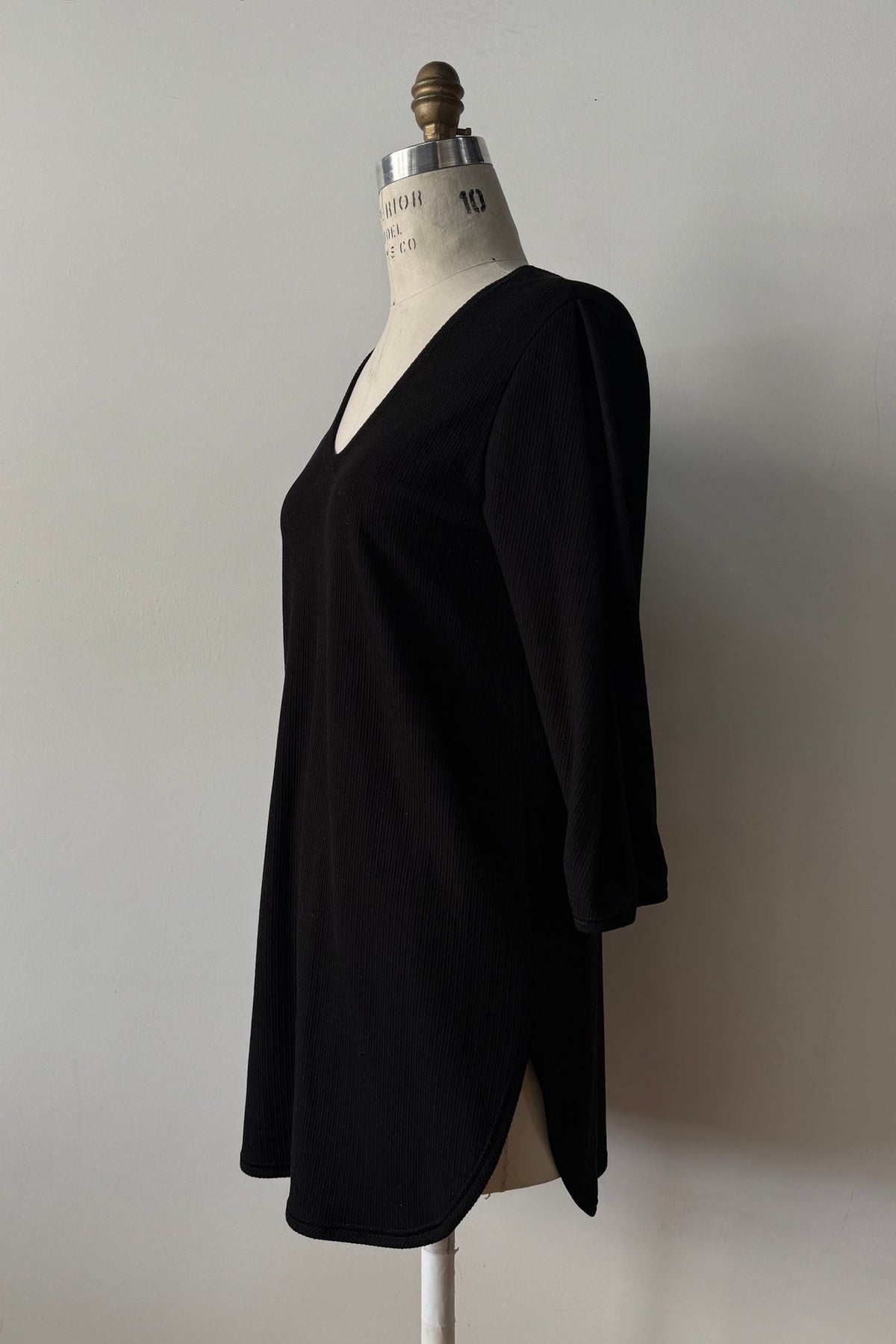 The Clara Tunic by Kollontai in Black, a stretch corduroy piece with a V-neck, a rounded hem, 3/4 sleeves, and inverted pleats at the shoulders, is shown on a mannequin against a grey background. 