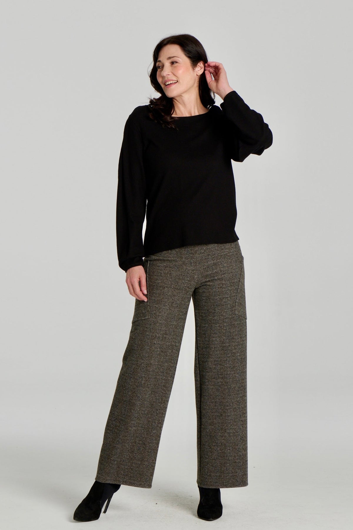 A woman wearing the Virginia Top by Kollontai in Black, a corduroy-textured top with inverted pleats on the long sleeves, a slim fit and a round neck. She is wearing it with the Susan pants in Chocolate and standing in front of a grey background. 