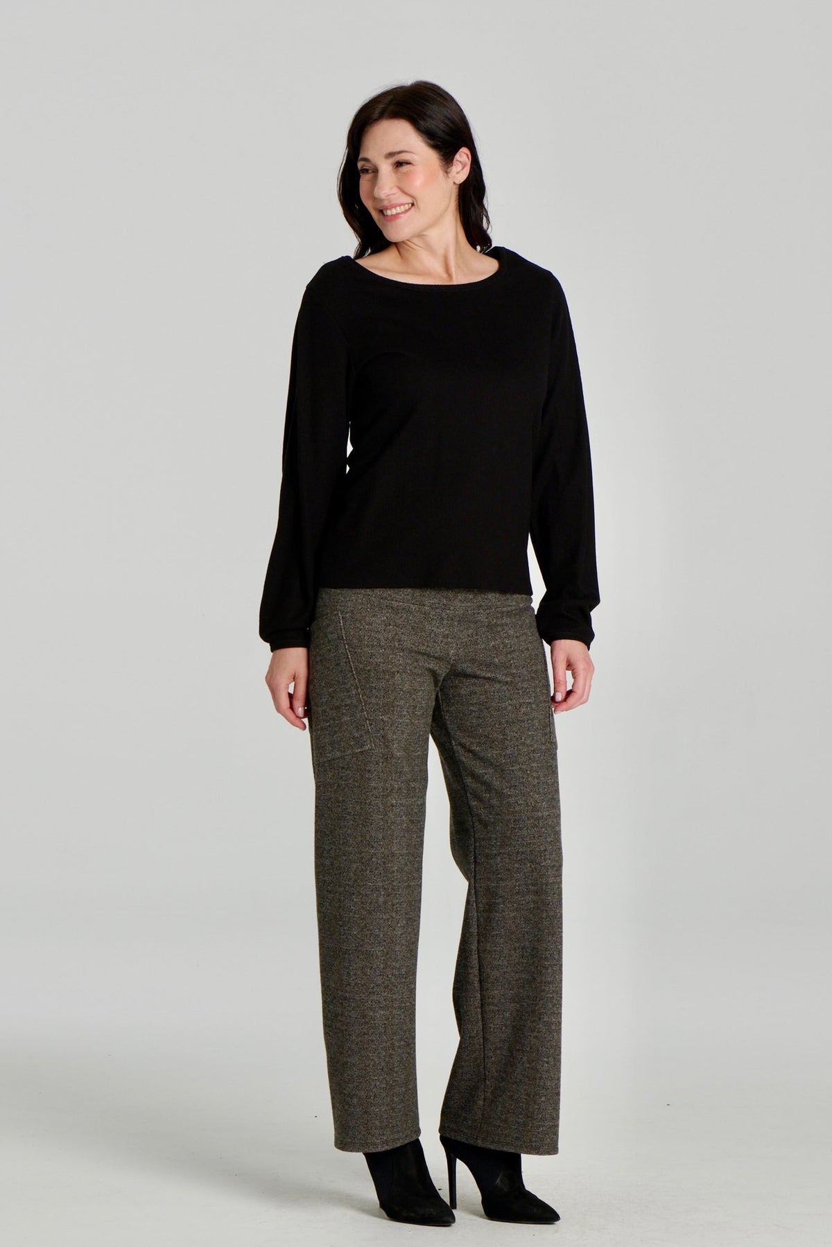 A woman wearing the Virginia Top by Kollontai in Black, a corduroy-textured top with inverted pleats on the long sleeves, a slim fit and a round neck. She is wearing it with the Susan pants in Chocolate and standing in front of a grey background. 