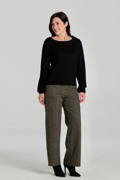 A woman wearing the Virginia Top by Kollontai in Black, a corduroy-textured top with inverted pleats on the long sleeves, a slim fit and a round neck. She is wearing it with the Susan pants in Chocolate and standing in front of a grey background. 