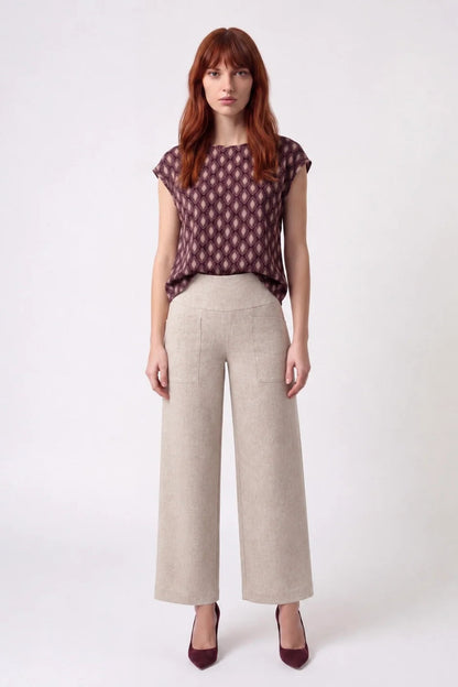 A woman wearing the Fanie Shirt by Kollontai, a relaxed fitting top with cape sleeves and a hi-low hem in a pink and plum geometric print. She is wearing it with cream pants and standing against a white background. 