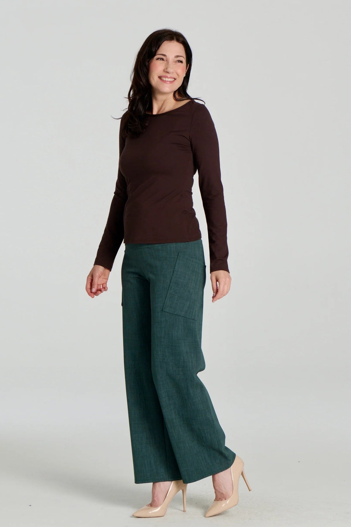 A woman wearing the Loretto Top by Kollontai in Chocolate, a slim fitting long sleeved top with a round neck. She is wearing it with the Susan pants in Green and standing in front of a grey background. 