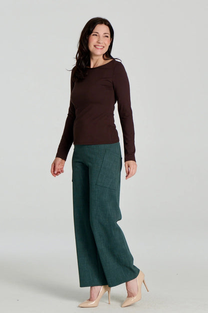 A woman wearing the Loretto Top by Kollontai in Chocolate, a slim fitting long sleeved top with a round neck. She is wearing it with the Susan pants in Green and standing in front of a grey background. 